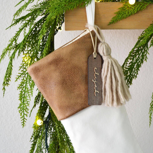 A brown leather Christmas stocking with a tassel and a wooden tag hanging from it, surrounded by greenery and twinkling lights.