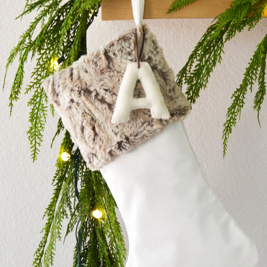 A white Christmas stocking decorated with a furry fabric and a letter "A" hanging from it, surrounded by greenery and twinkling lights.