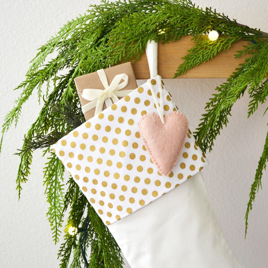 A white Christmas stocking adorned with a gold polka dot pattern, a pink felt heart, and a small gift box tied with a white ribbon. The stocking is hanging on a wooden hanger, and a garland of pine branches is draped around it, creating a festive and cozy holiday atmosphere.