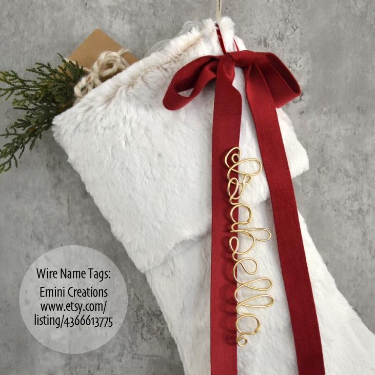 A white fur stocking with a red ribbon and a personalized name tag attached to it, hanging on a gray wall.