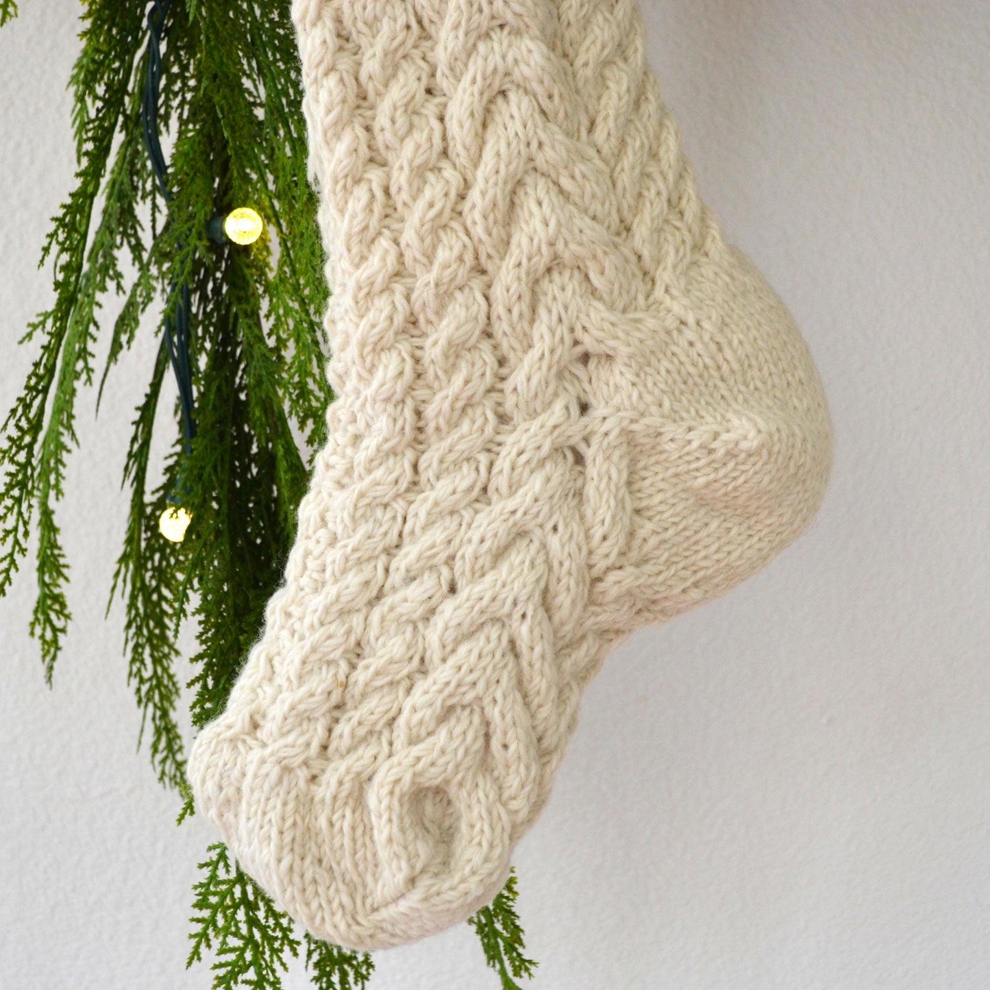 A close-up view of a white knitted stocking with intricate braided patterns, hanging from a branch with green pine needles and twinkling lights.