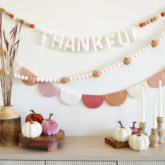 a festive Thanksgiving display with a banner that says "THANKFUL" hanging on a white wall. The display includes a wooden table with a vase of pumpkins, candles, and other decorative items.