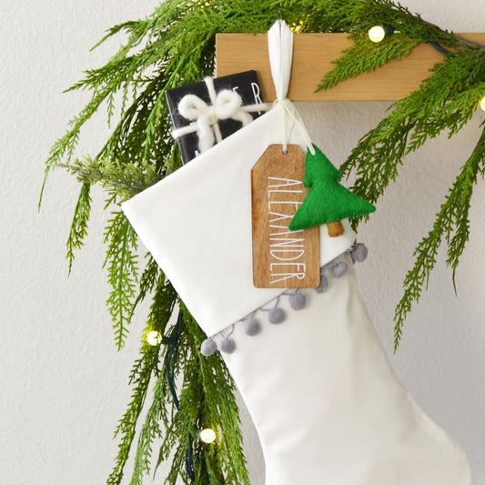 A white Christmas stocking decorated with greenery, a wooden tag, and a small green ornament, hanging on a wooden hanger.
