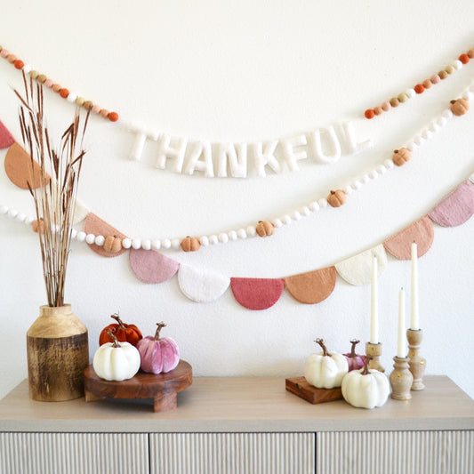 a festive Thanksgiving display with a string of colorful beads hanging on a wall, a wooden table with a vase of pumpkins, and a banner that says "THANKFUL".