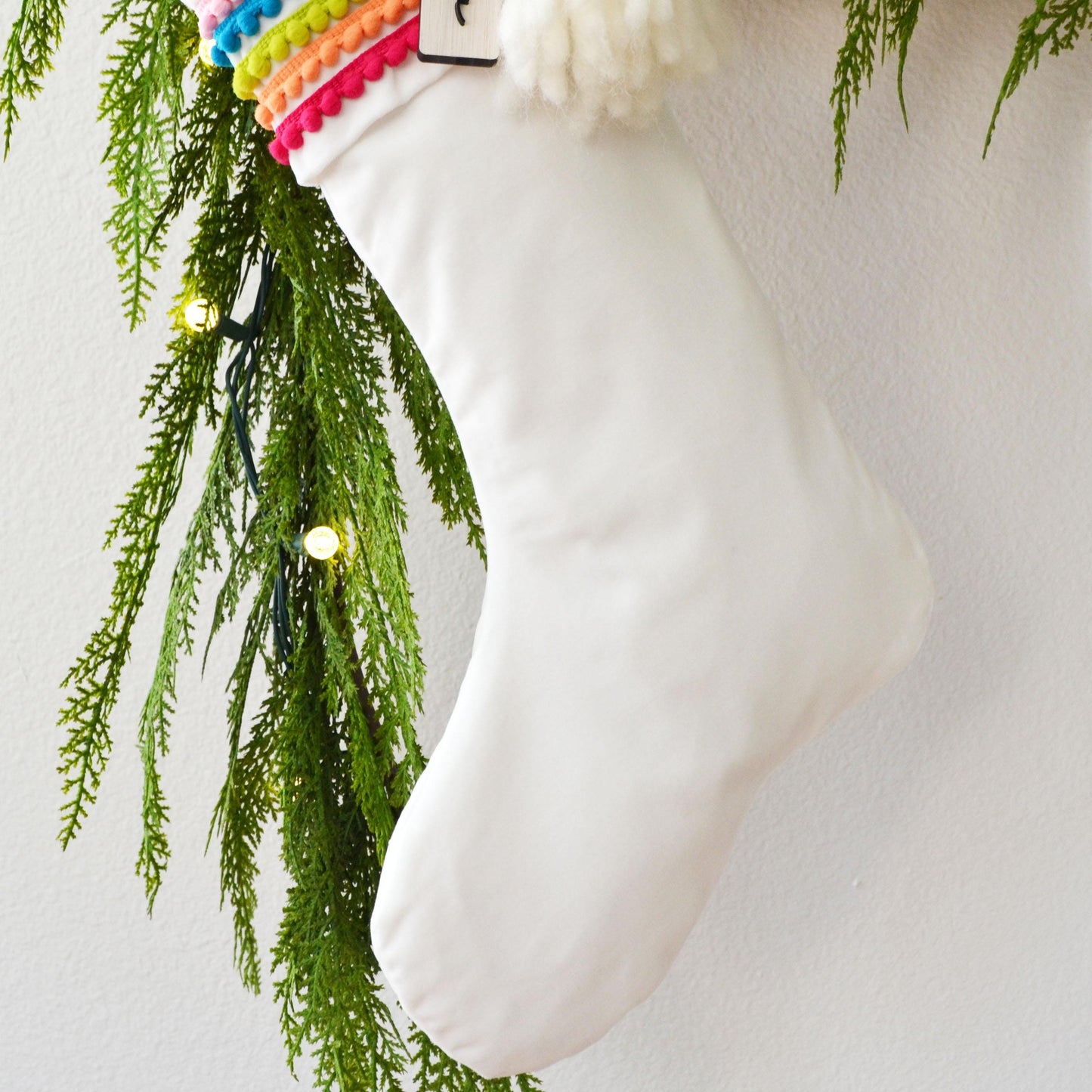 A white Christmas stocking adorned with colorful beads and greenery, hanging on a wall.
