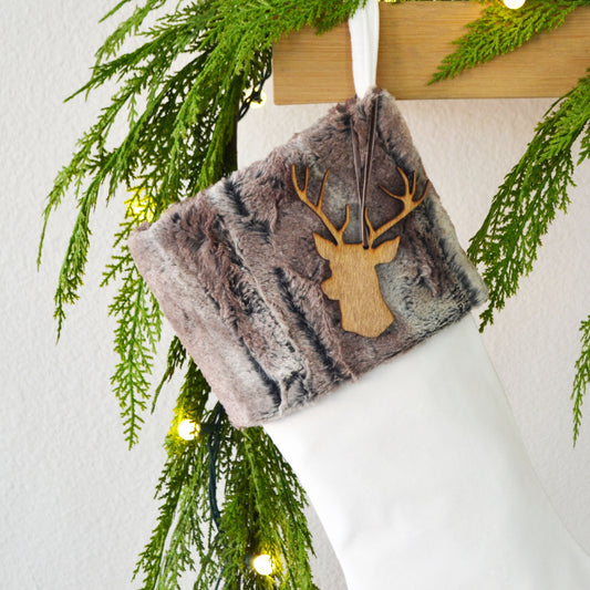 A white Christmas stocking adorned with a brown and gray fur-like material and a wooden deer head ornament, hanging on a wooden hanger.