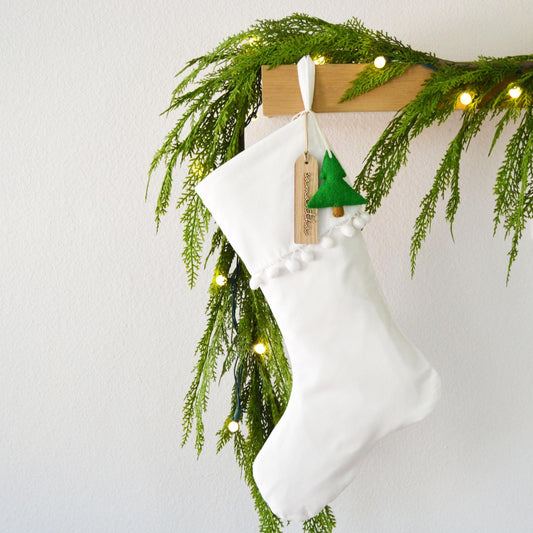 A white Christmas stocking hangs on a wooden hanger, adorned with a garland of greenery and twinkling lights.