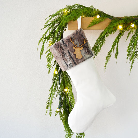 A white Christmas stocking adorned with a brown deer head and a fur trim hangs from a wooden hanger, decorated with a garland of greenery and twinkling lights.