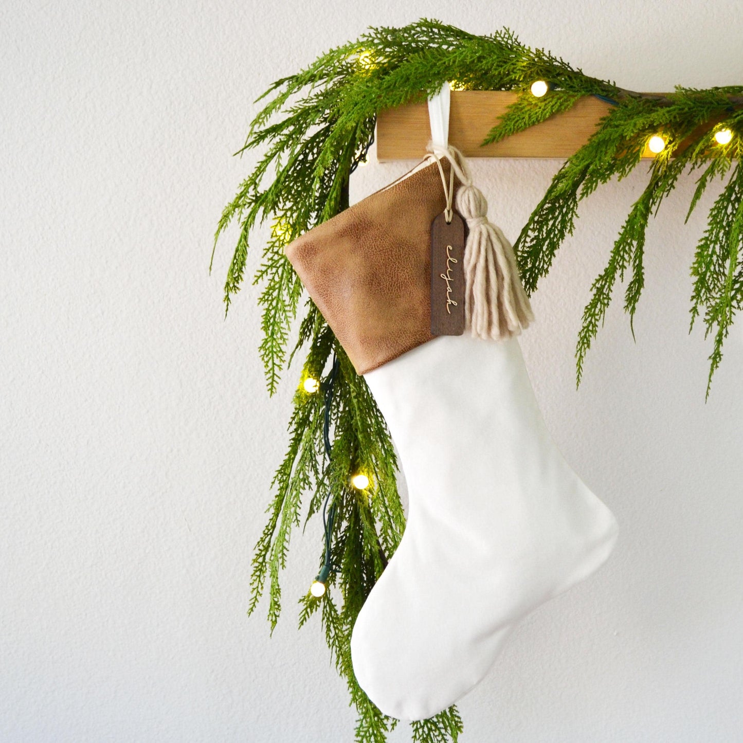 A white Christmas stocking adorned with greenery and twinkling lights, hanging on a wooden hanger.