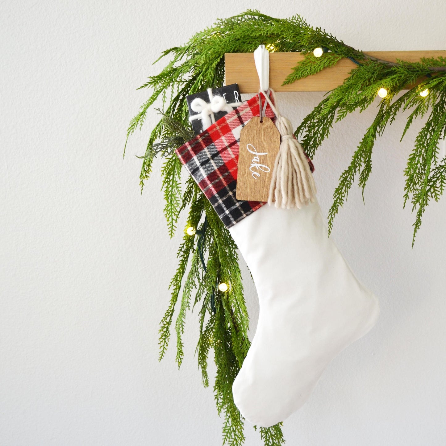 A white Christmas stocking adorned with a red and black plaid blanket, a tassel, and a small gift tag hangs from a wooden hanger. The stocking is decorated with greenery and twinkling lights, creating a festive atmosphere.