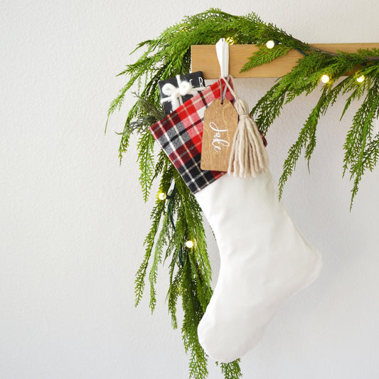 A white Christmas stocking adorned with a red and black plaid blanket, a tassel, and a small gift tag hangs from a wooden hanger. The stocking is decorated with greenery and twinkling lights, creating a festive atmosphere.