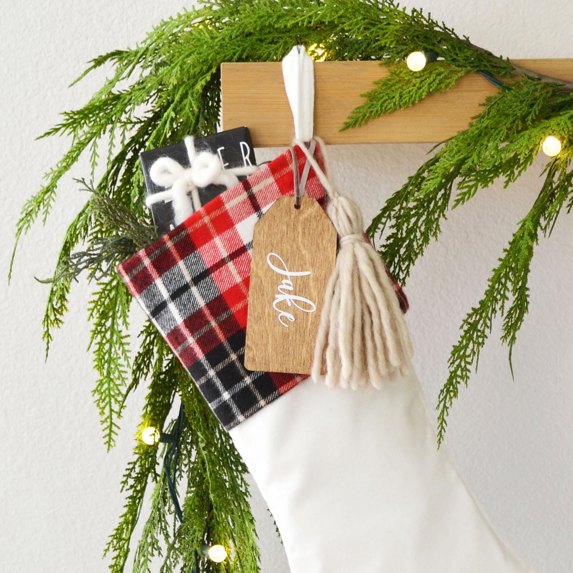A white Christmas stocking decorated with a red and black plaid fabric, a wooden tag with the name "Jale", and a white tassel. The stocking is hanging on a wooden hanger, and there are green pine branches and twinkling lights in the background.