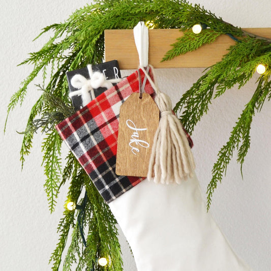 A white Christmas stocking decorated with a red and black plaid fabric, a wooden tag with the name "Jale", and a white tassel. The stocking is hanging on a wooden hanger, and there are green pine branches and twinkling lights in the background.