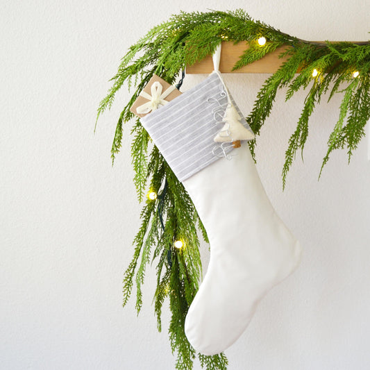 A white Christmas stocking hangs on a wall, adorned with a garland of greenery and twinkling lights.