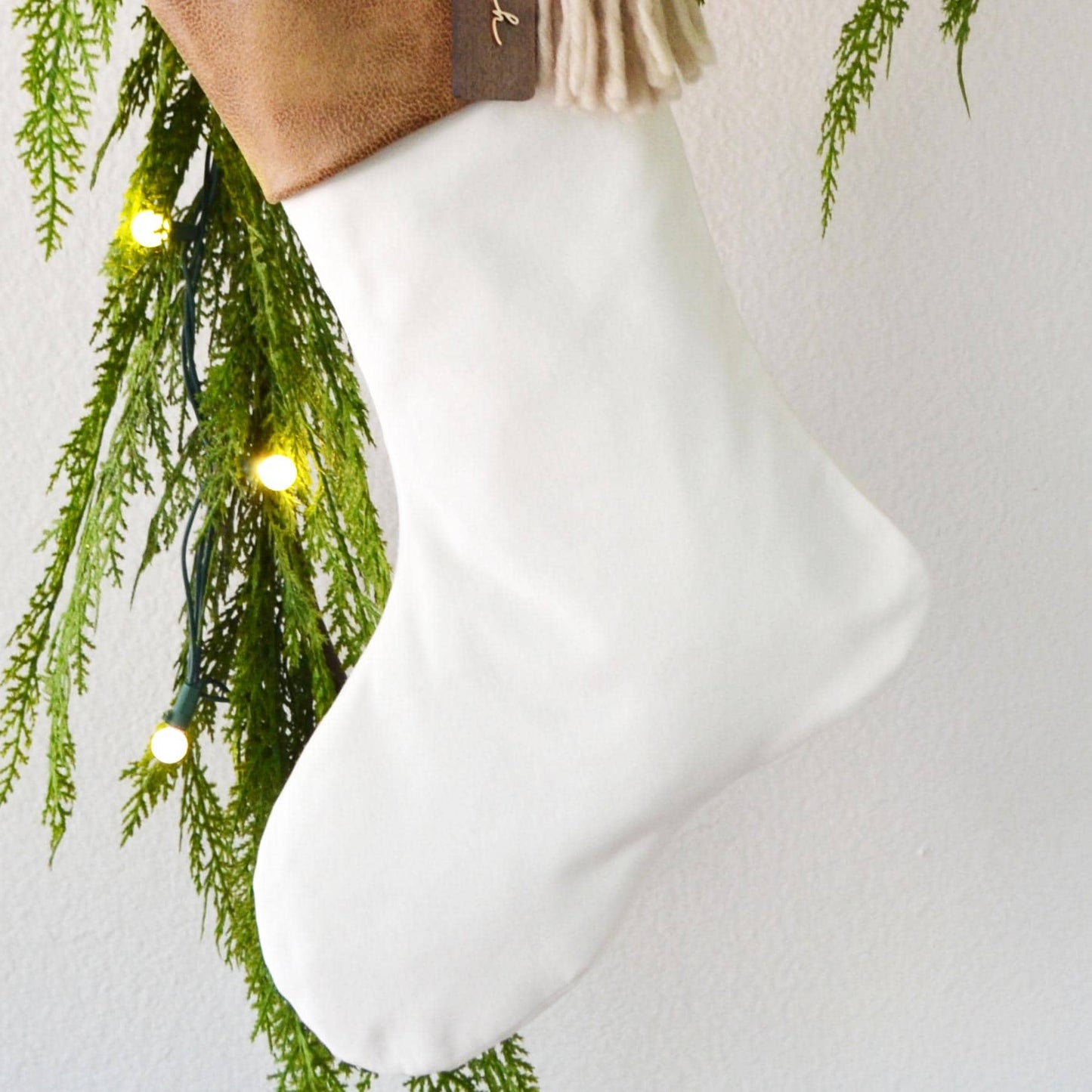 A white Christmas stocking is hanging from a branch with green pine needles and twinkling lights.