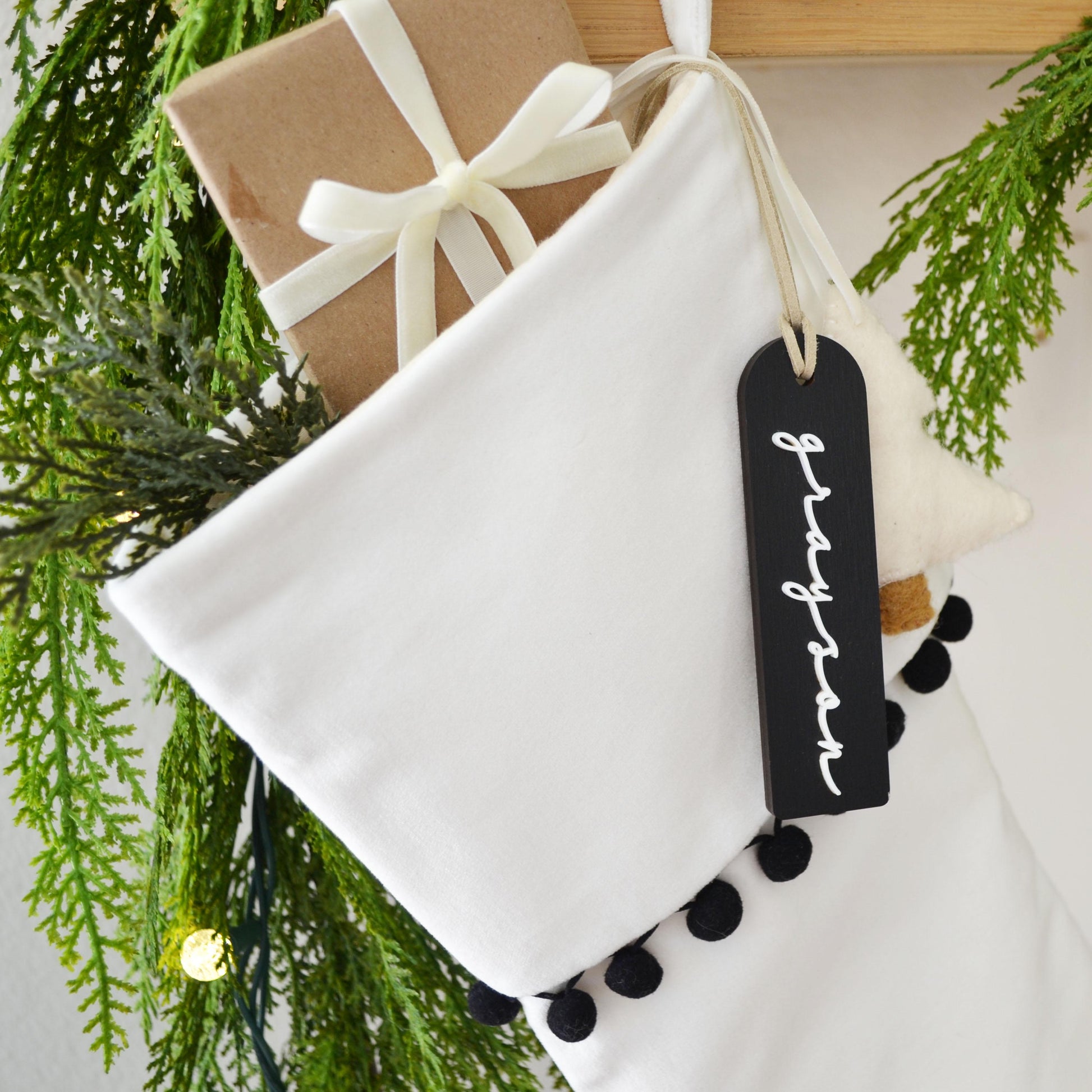 A white Christmas stocking decorated with pine branches, a gift box, and a black tag with the name "Grayson" written on it.