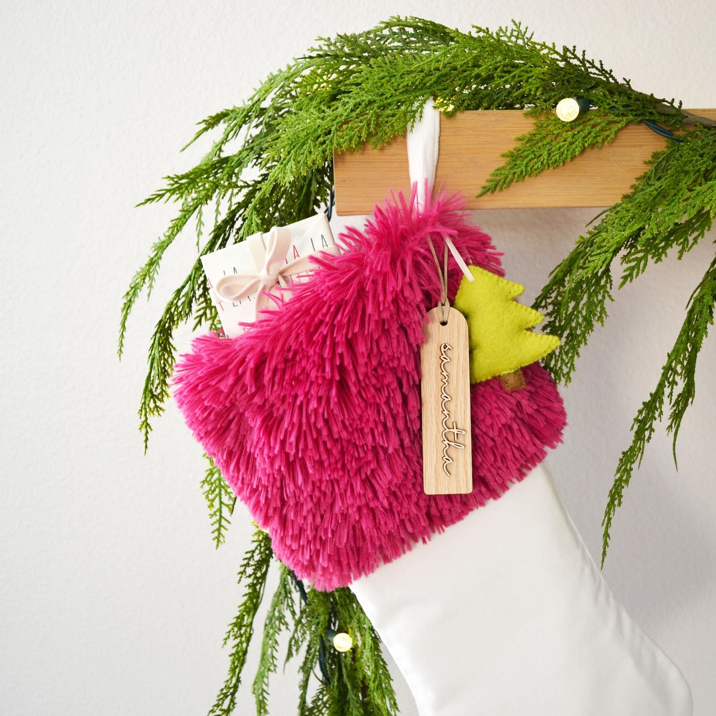 A pink furry stocking with a yellow tree ornament and a white tag hangs on a wooden hanger, surrounded by greenery and twinkling lights.
