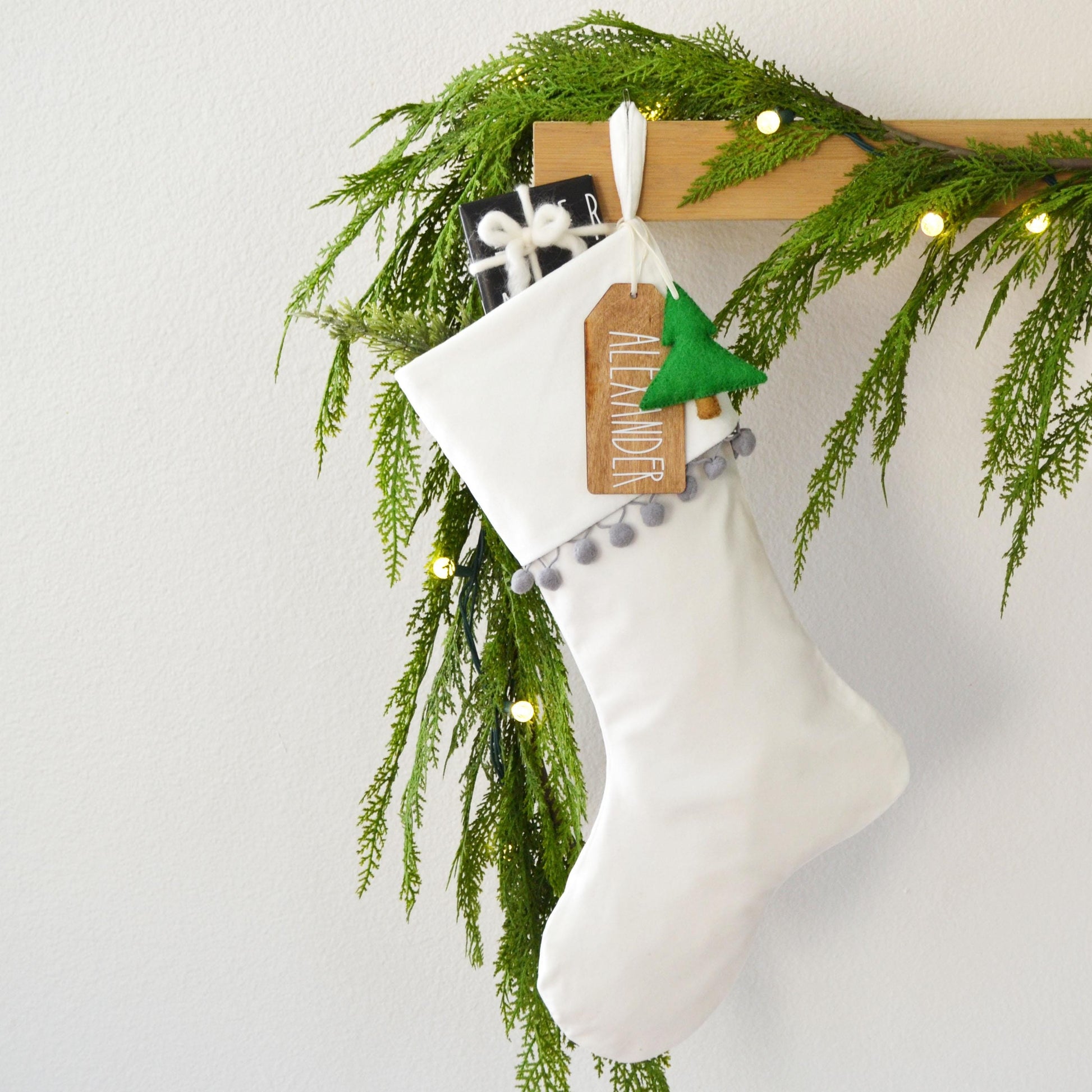 A white Christmas stocking decorated with greenery and a small green ornament, hanging on a wooden hanger.