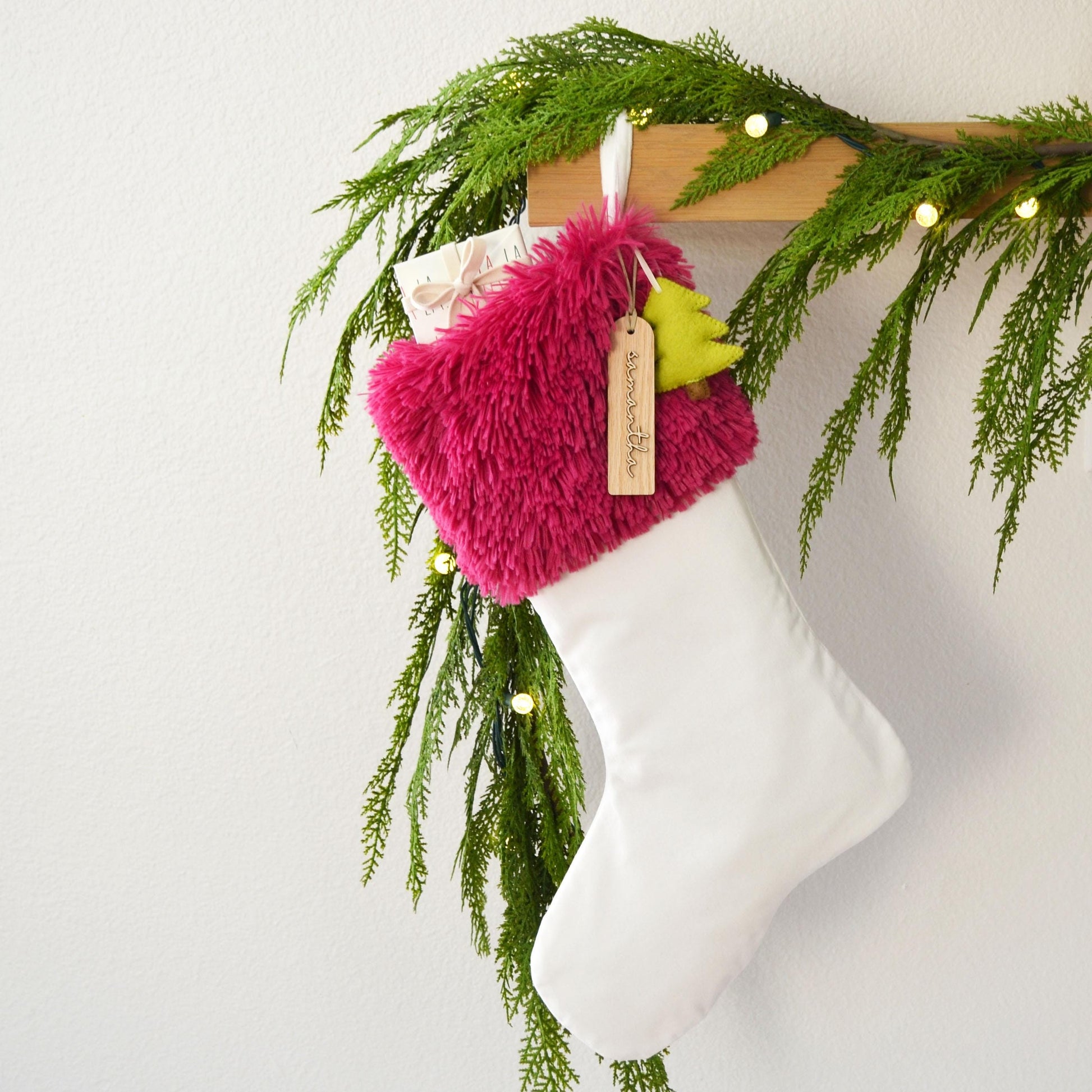 A white Christmas stocking with a pink furry trim and a yellow tag is hanging on a wooden hanger, decorated with greenery and twinkling lights.