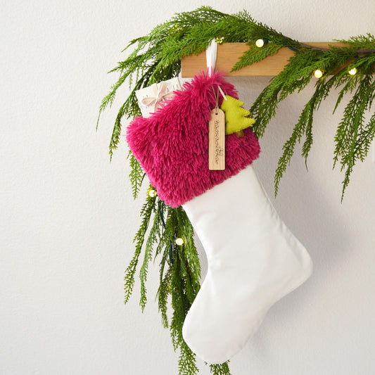 A white Christmas stocking with a pink furry trim and a yellow tag is hanging on a wooden hanger, decorated with greenery and twinkling lights.