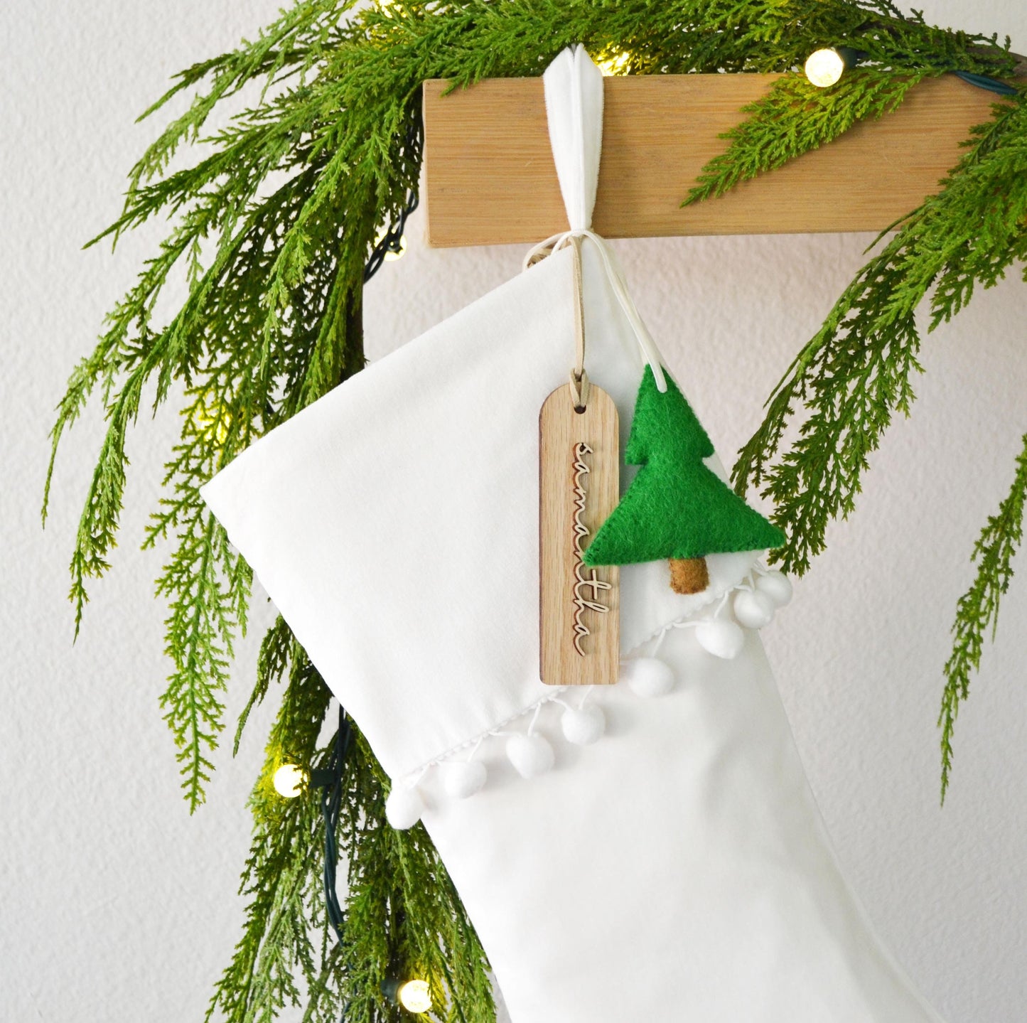 A white Christmas stocking adorned with a green felt tree ornament and a wooden tag hangs on a wooden hanger, surrounded by a garland of pine needles and twinkling lights.