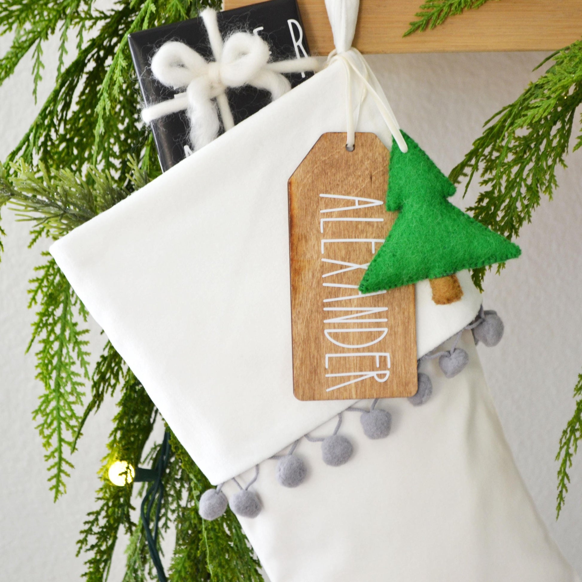 A white Christmas stocking decorated with a green felt tree, a wooden tag with the name "ALEXANDER", and gray pom-poms.
