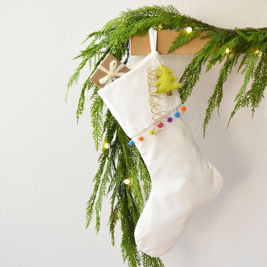 A white Christmas stocking adorned with festive decorations, including a green tree ornament and colorful balls, hangs from a wooden hanger against a white wall.