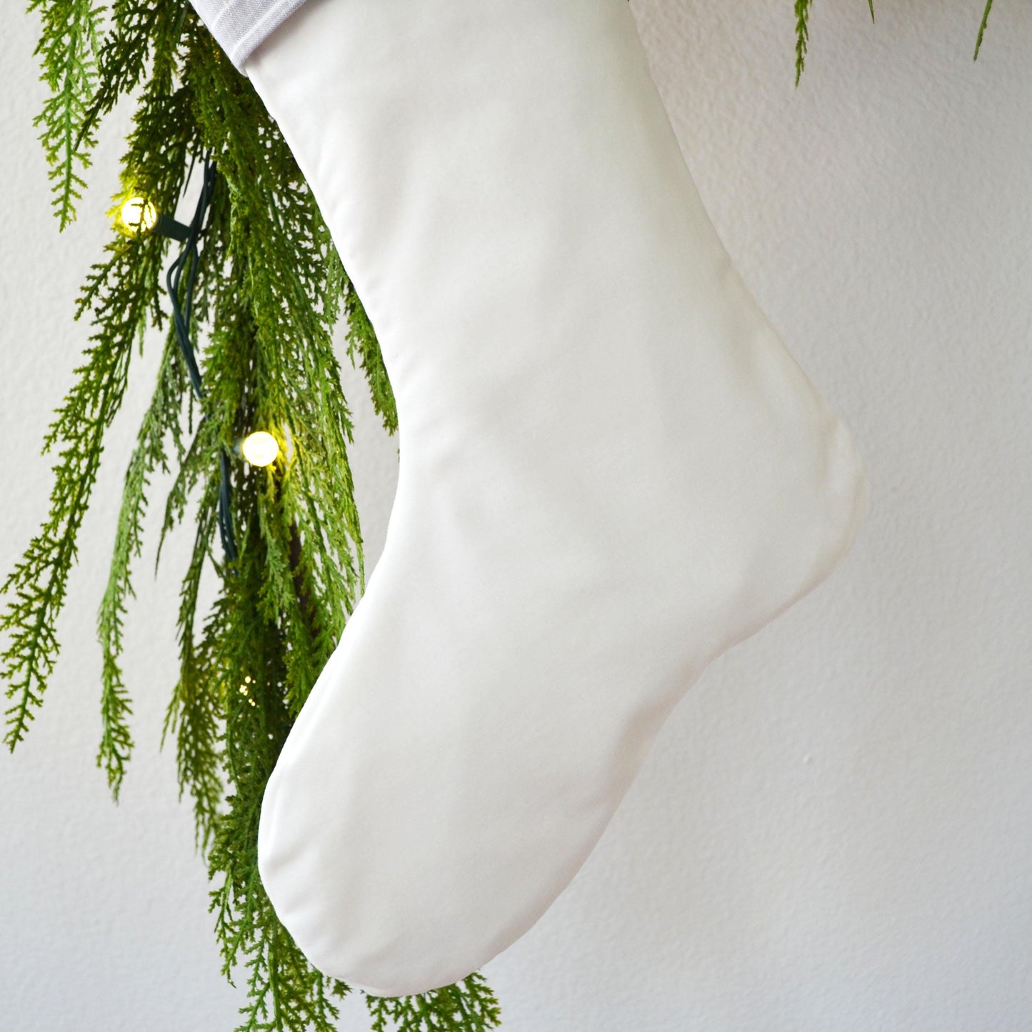 A white Christmas stocking is hanging from a tree branch with green pine needles and twinkling lights.