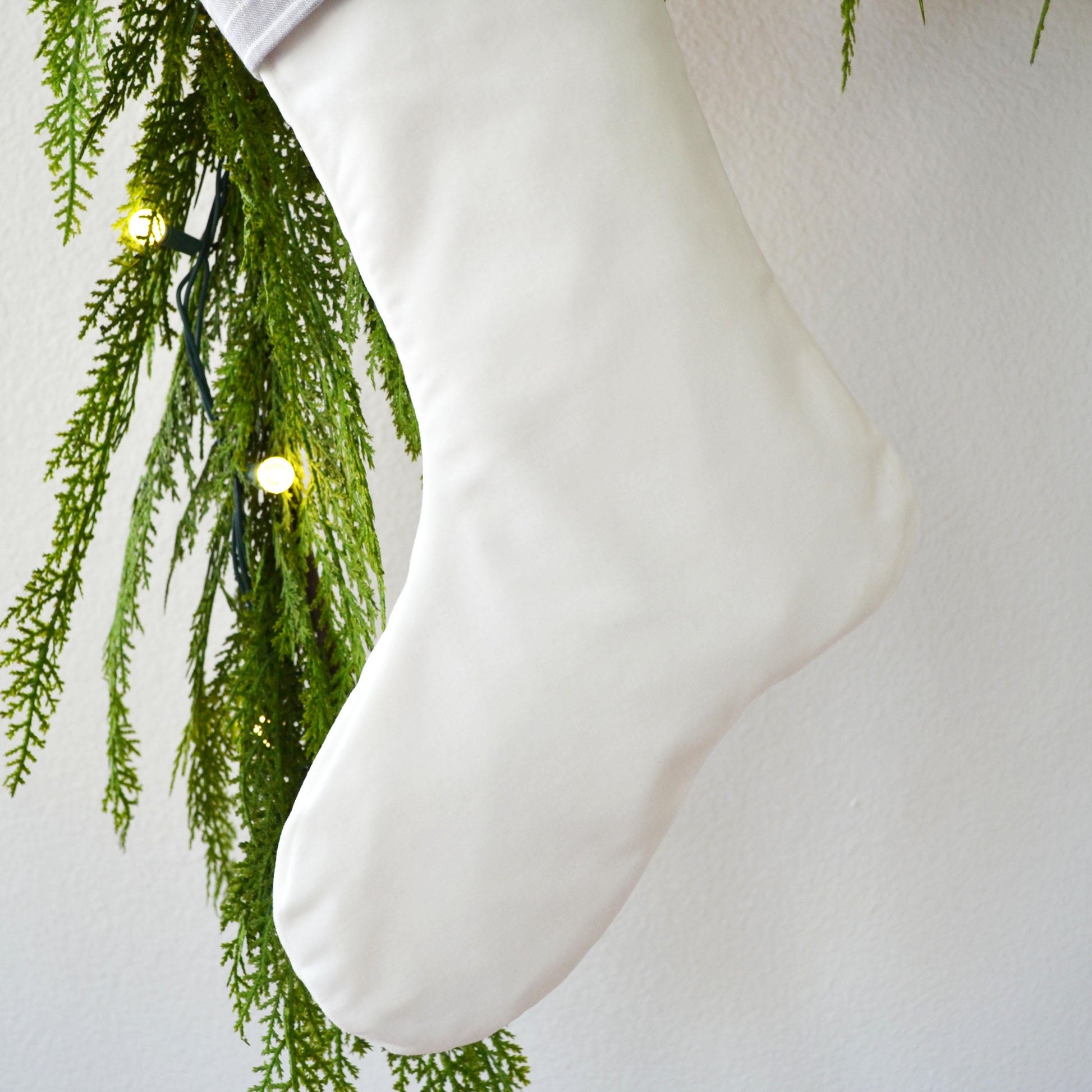 A white Christmas stocking is hanging from a tree branch with green pine needles and twinkling lights.