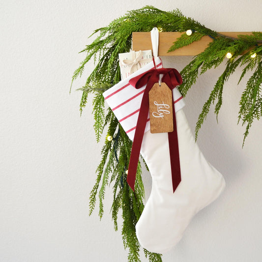 A white Christmas stocking adorned with greenery and a red ribbon, with a wooden tag attached that says "Lily".