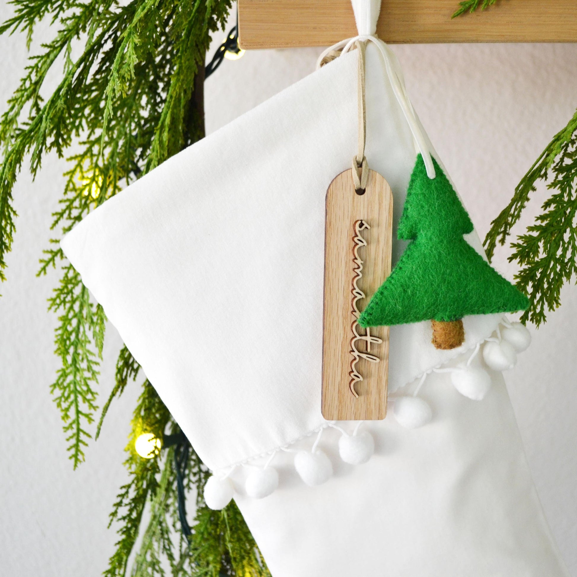 A white Christmas stocking decorated with a green felt tree ornament and a wooden tag with the name "Smitha" written on it, hanging on a wooden hanger.