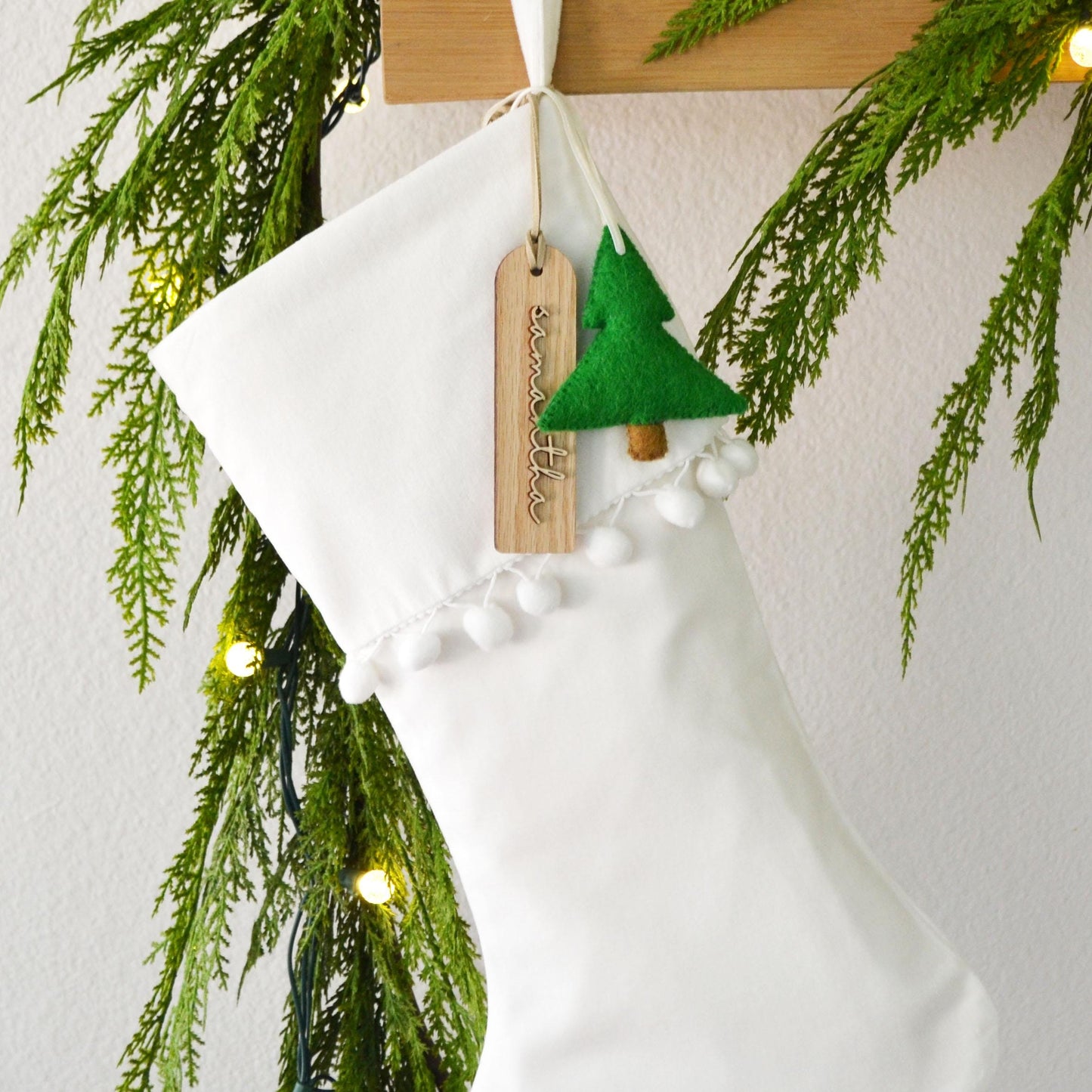 A white Christmas stocking decorated with a green felt tree and white pom-poms, hanging on a wooden hanger.