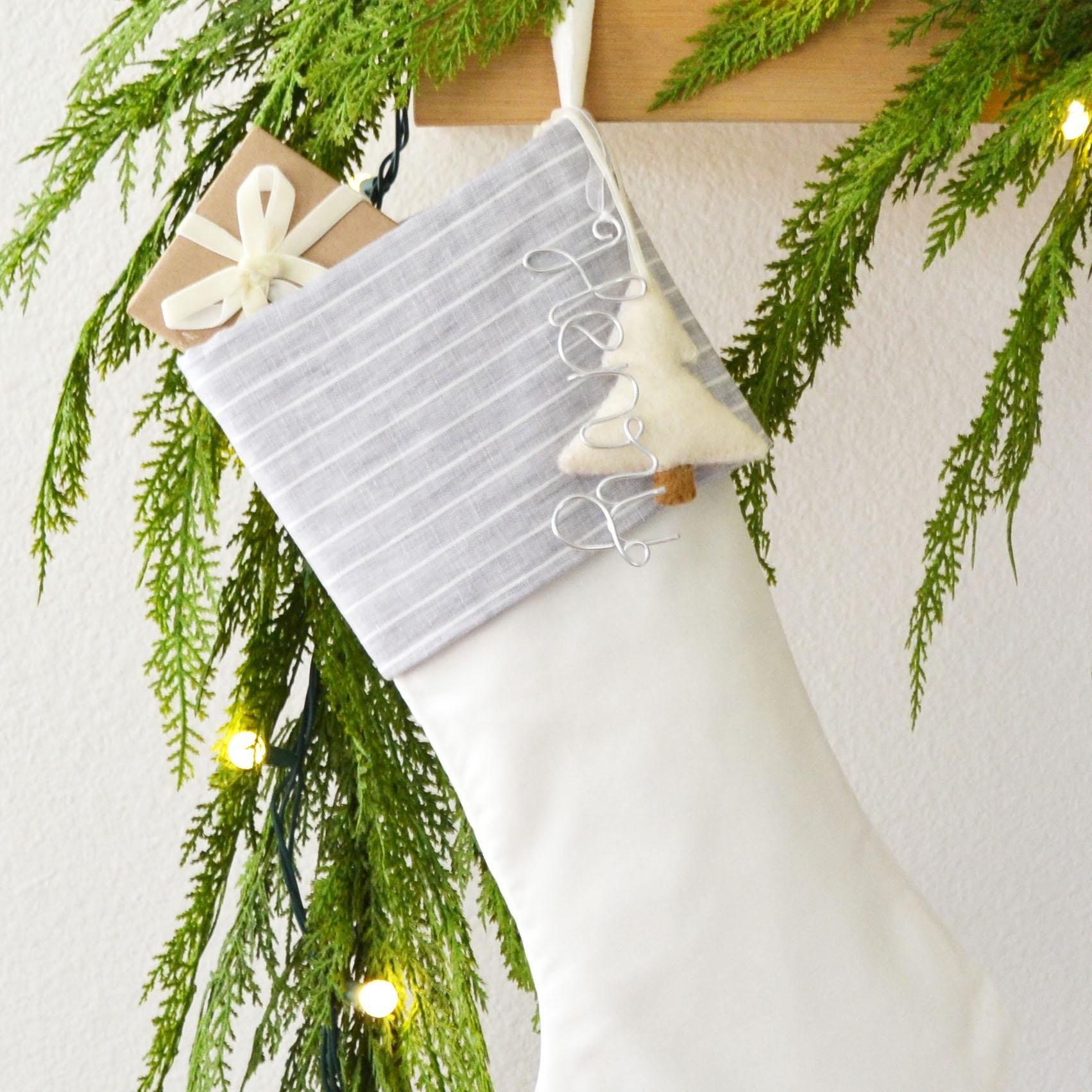 A white Christmas stocking decorated with a ribbon, a small white ornament, and greenery, hanging on a wooden hanger.