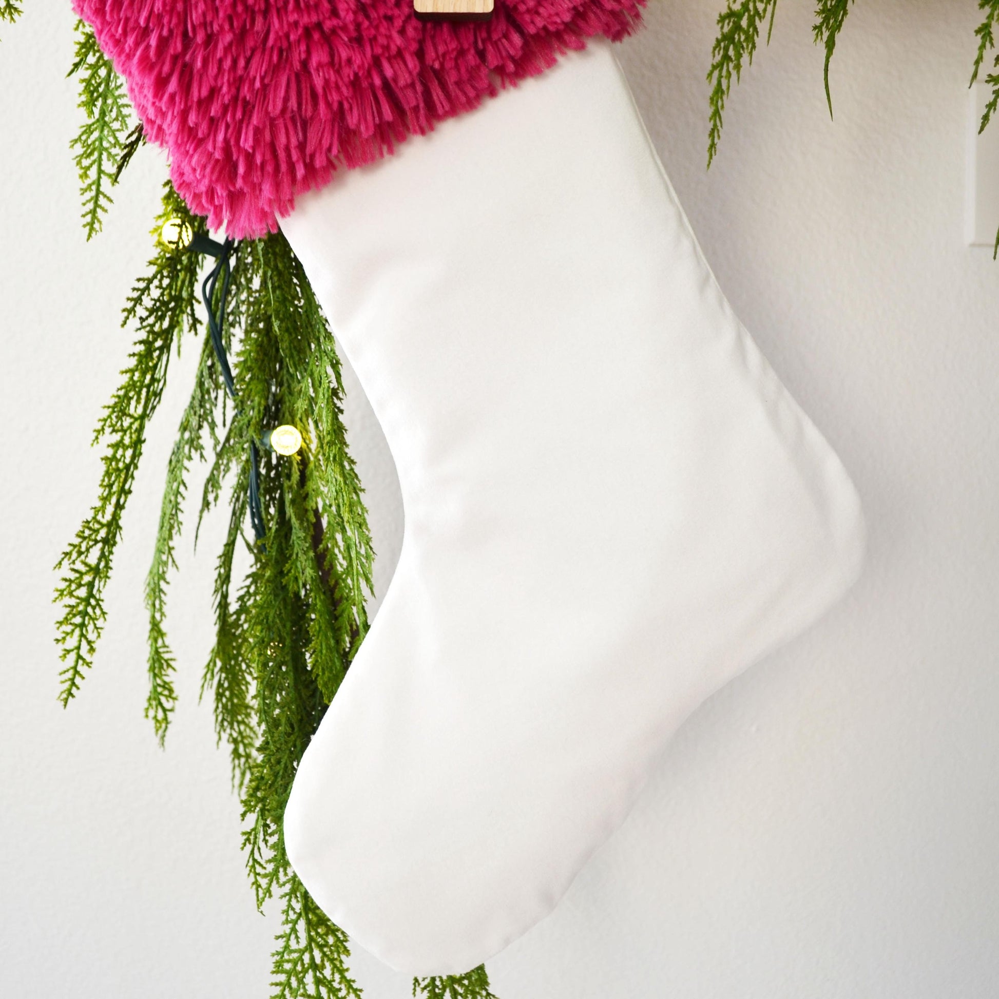 A white Christmas stocking with a pink fuzzy trim is hanging on a wall, decorated with greenery and twinkling lights.