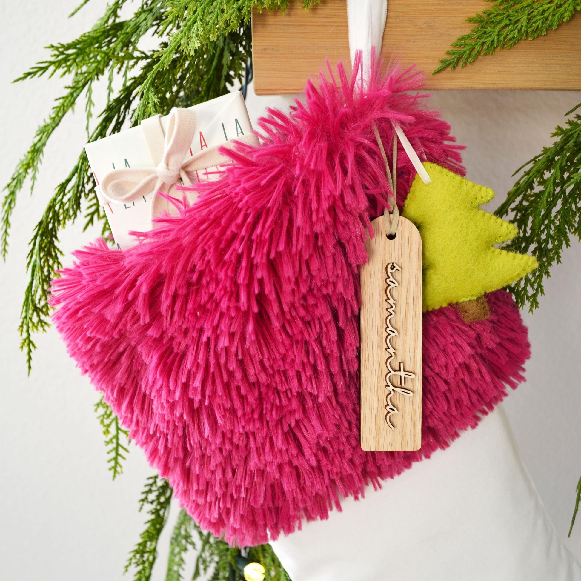 A pink furry stocking with a wooden tag and a yellow felt Christmas tree ornament are hanging on a wooden hanger, surrounded by greenery.