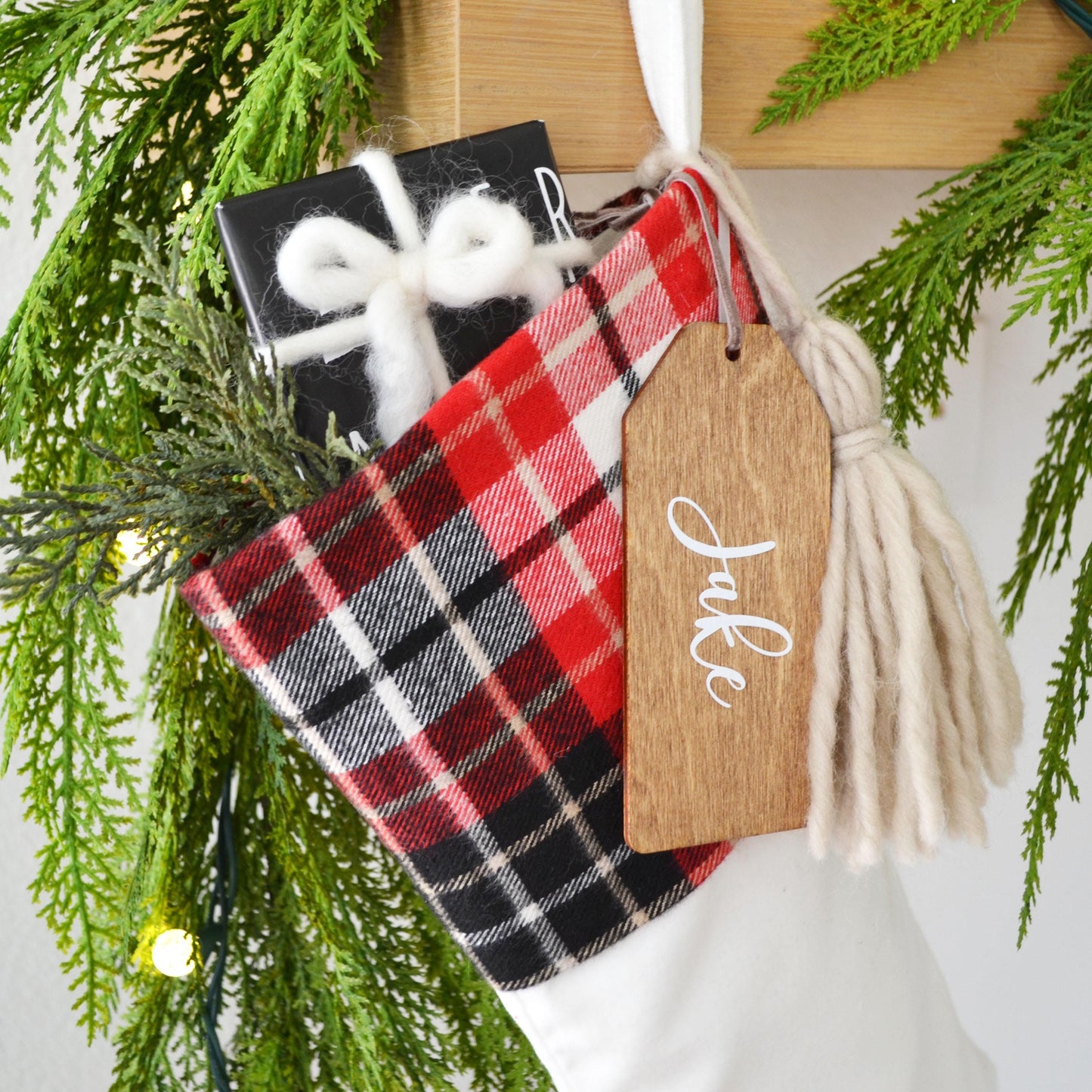 A Christmas stocking decorated with a red and black plaid fabric, a black gift box, and a wooden tag with the name "Jake" written on it, all set against a backdrop of greenery and twinkling lights.