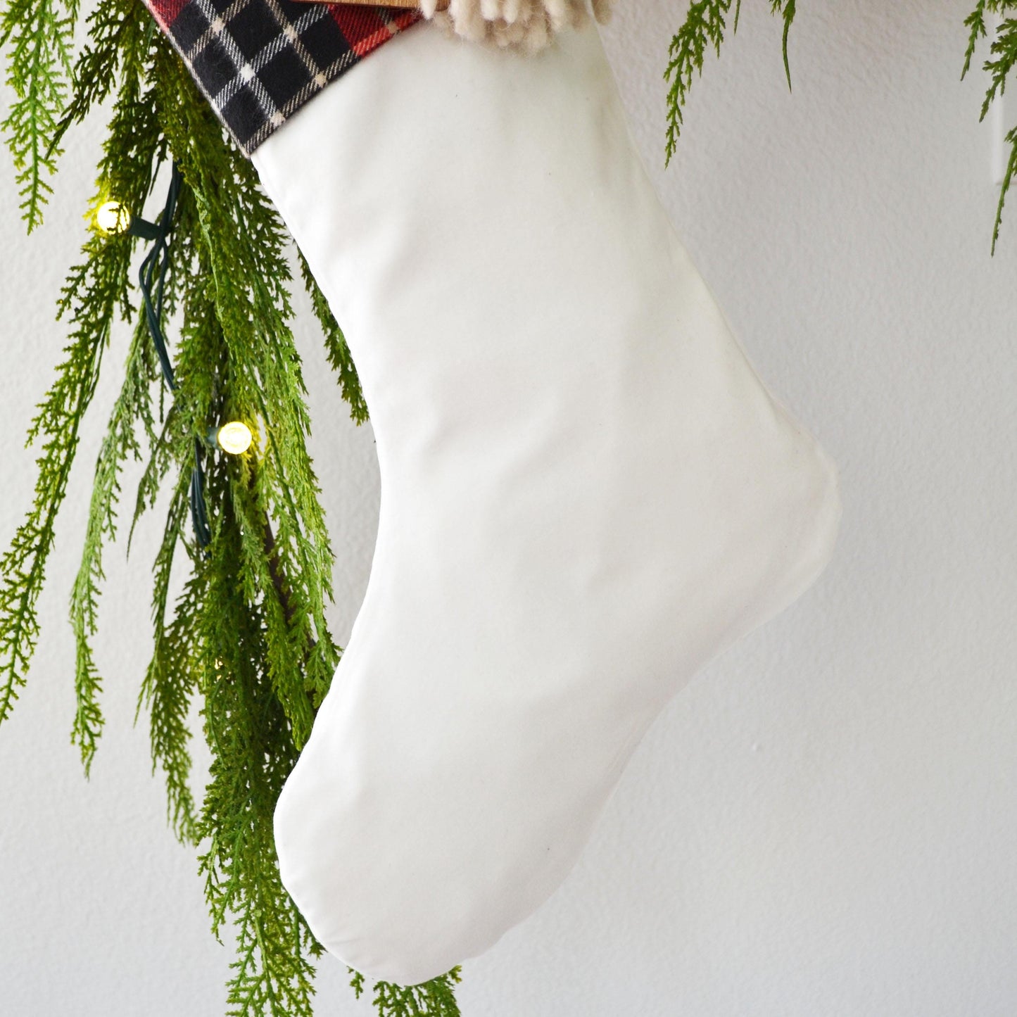 A white Christmas stocking is hanging from a tree branch with green pine needles and twinkling lights.