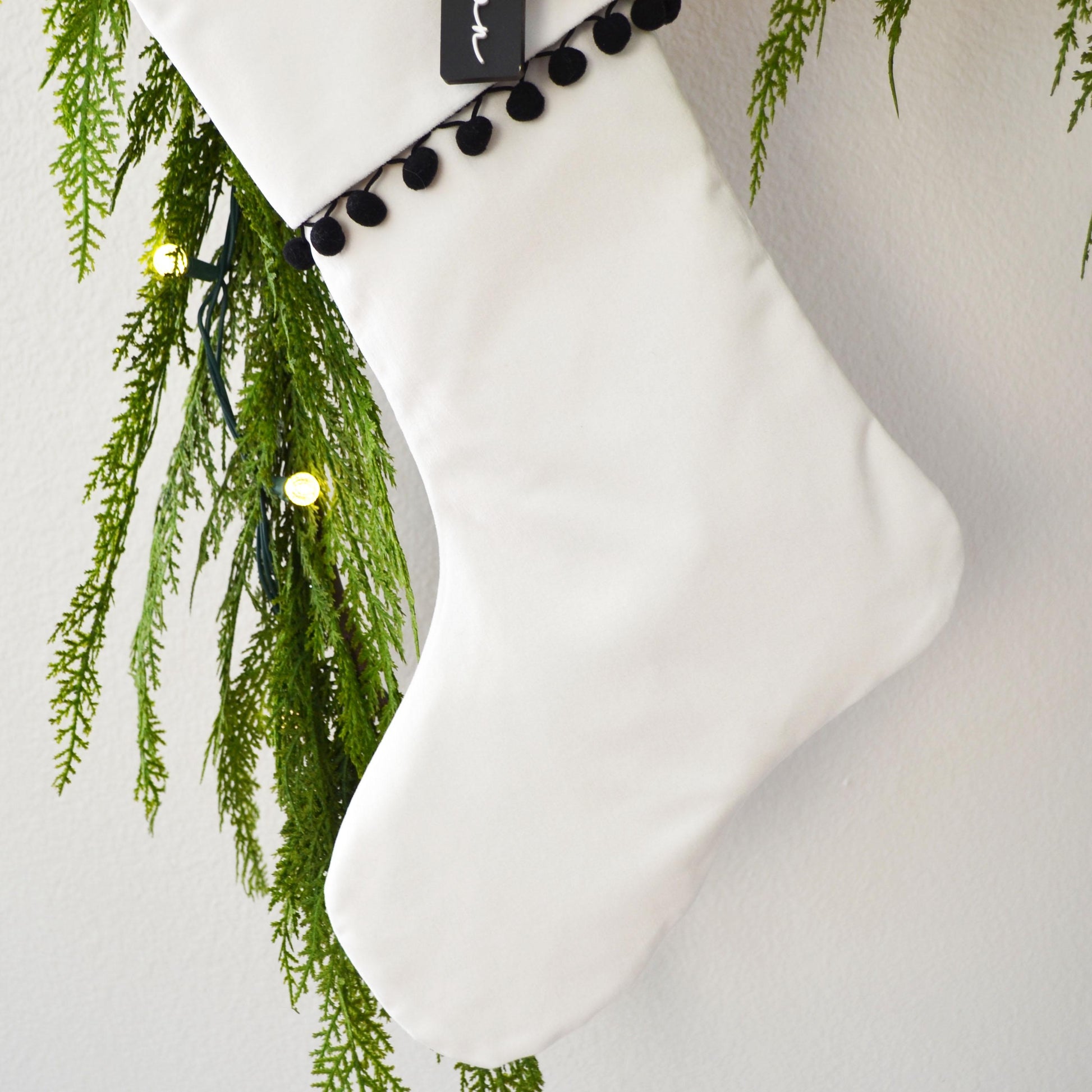 A white Christmas stocking adorned with greenery and twinkling lights, hanging against a white background.