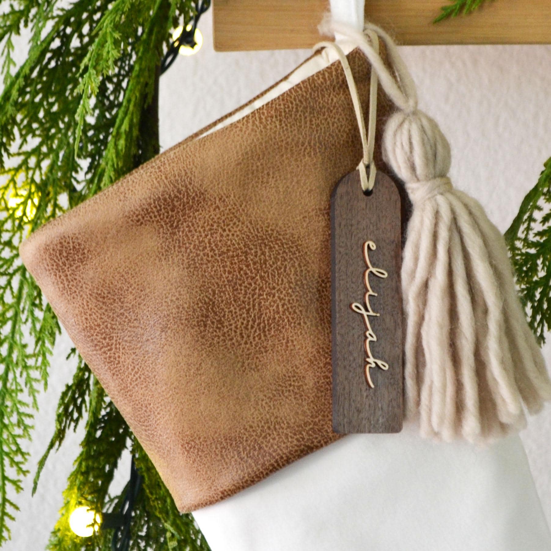 A brown leather-like material is hanging from a wooden hanger, with a wooden tag attached to it. The tag has the name "Logan" written on it in cursive script.