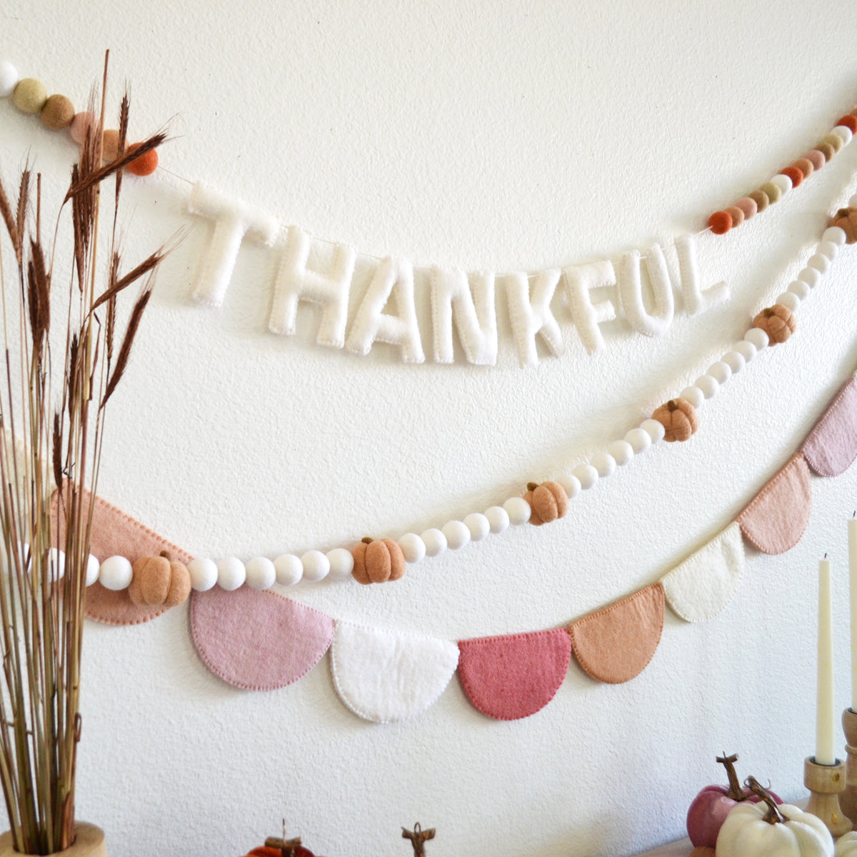 A decorative string of felt letters spelling "THANKFUL" hangs on a white wall, accompanied by a garland of beads and pumpkins.
