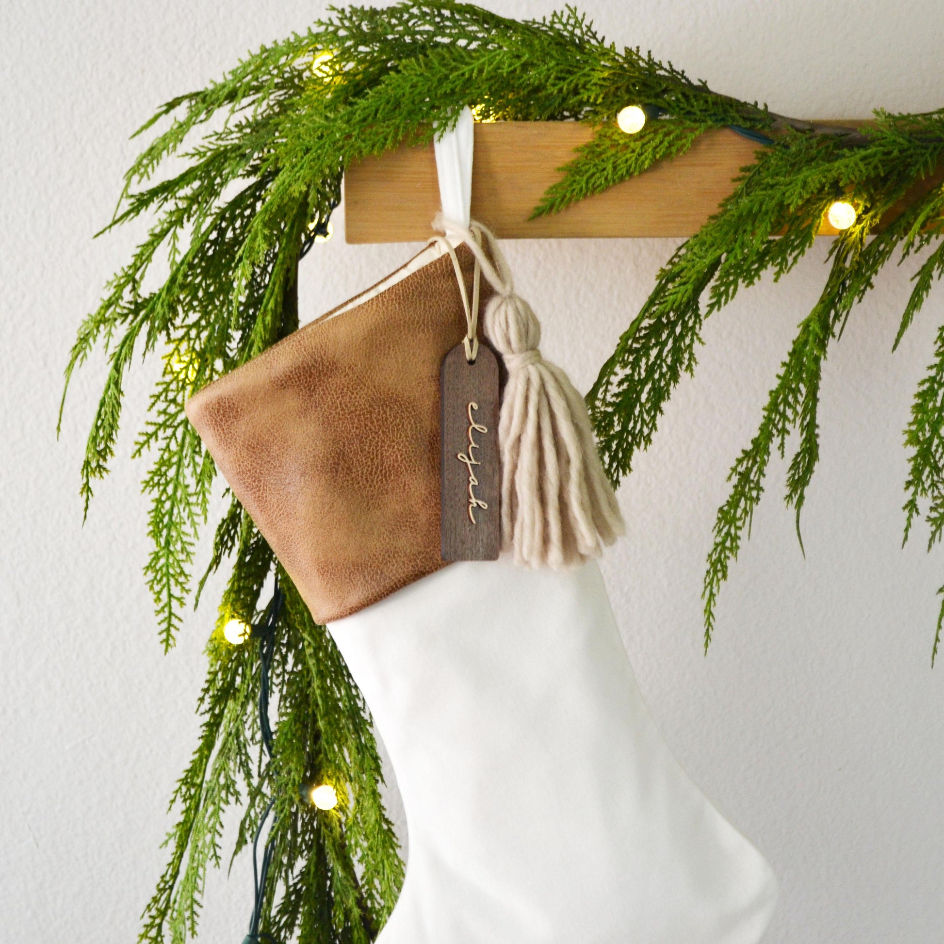A brown leather Christmas stocking with a tassel hangs on a wooden hanger, adorned with greenery and twinkling lights.