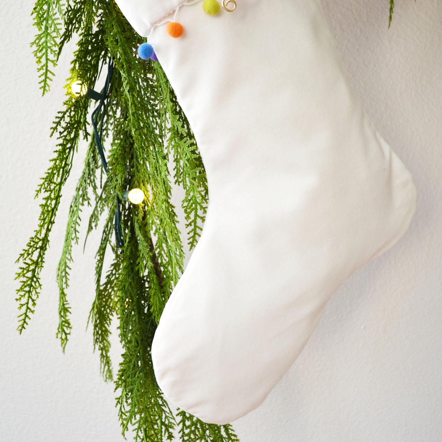 A white Christmas stocking adorned with greenery and colorful ornaments is hanging against a white wall.