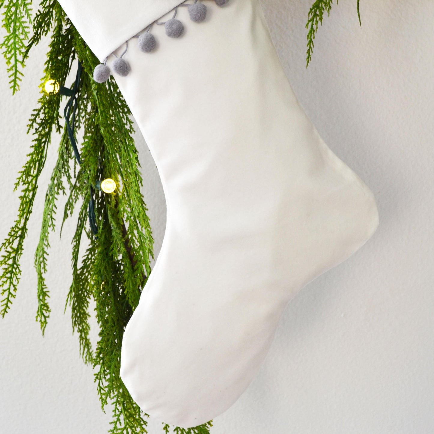 A white Christmas stocking adorned with greenery and small white balls hangs against a white wall.