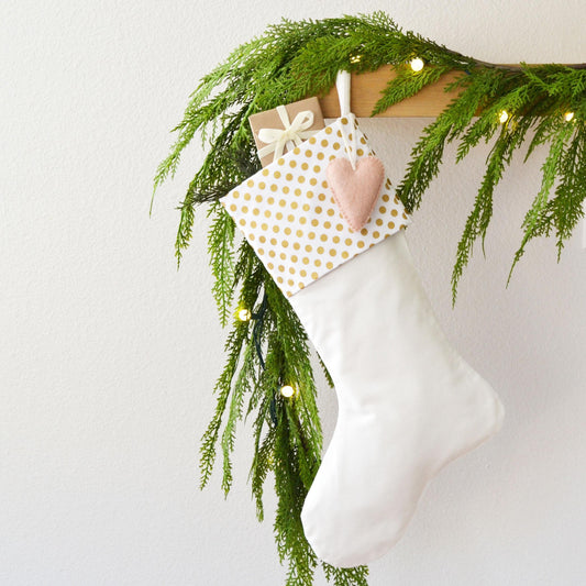 A white Christmas stocking adorned with a polka dot pattern and a pink heart ornament, hanging from a branch of greenery and twinkling lights.