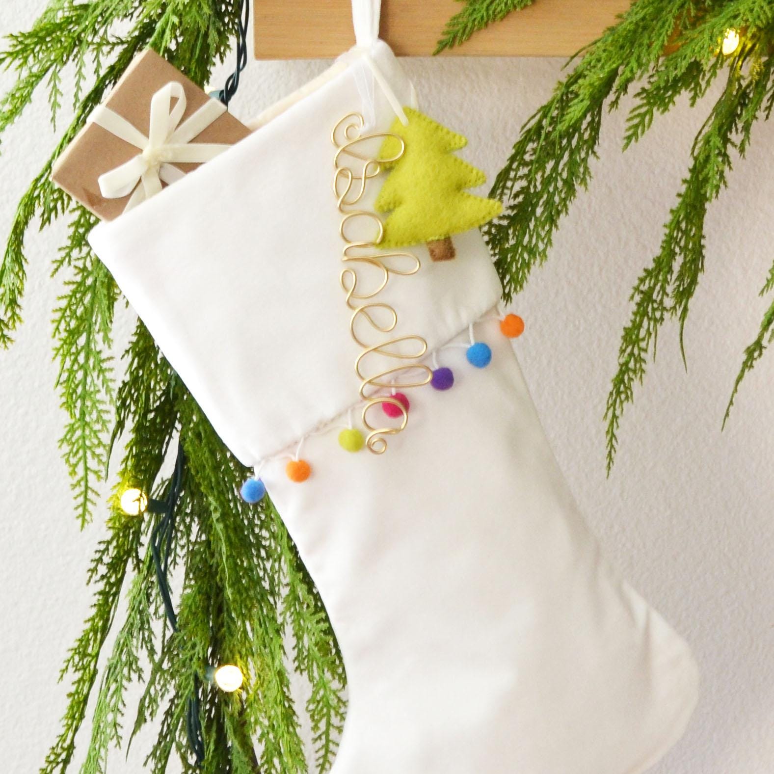 A white Christmas stocking decorated with a yellow tree ornament, colorful balls, and a gift box, hanging on a wall with greenery and twinkling lights.