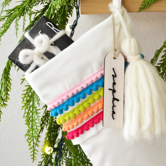 A white Christmas stocking decorated with colorful pom poms and a tassel, hanging on a wooden hanger.