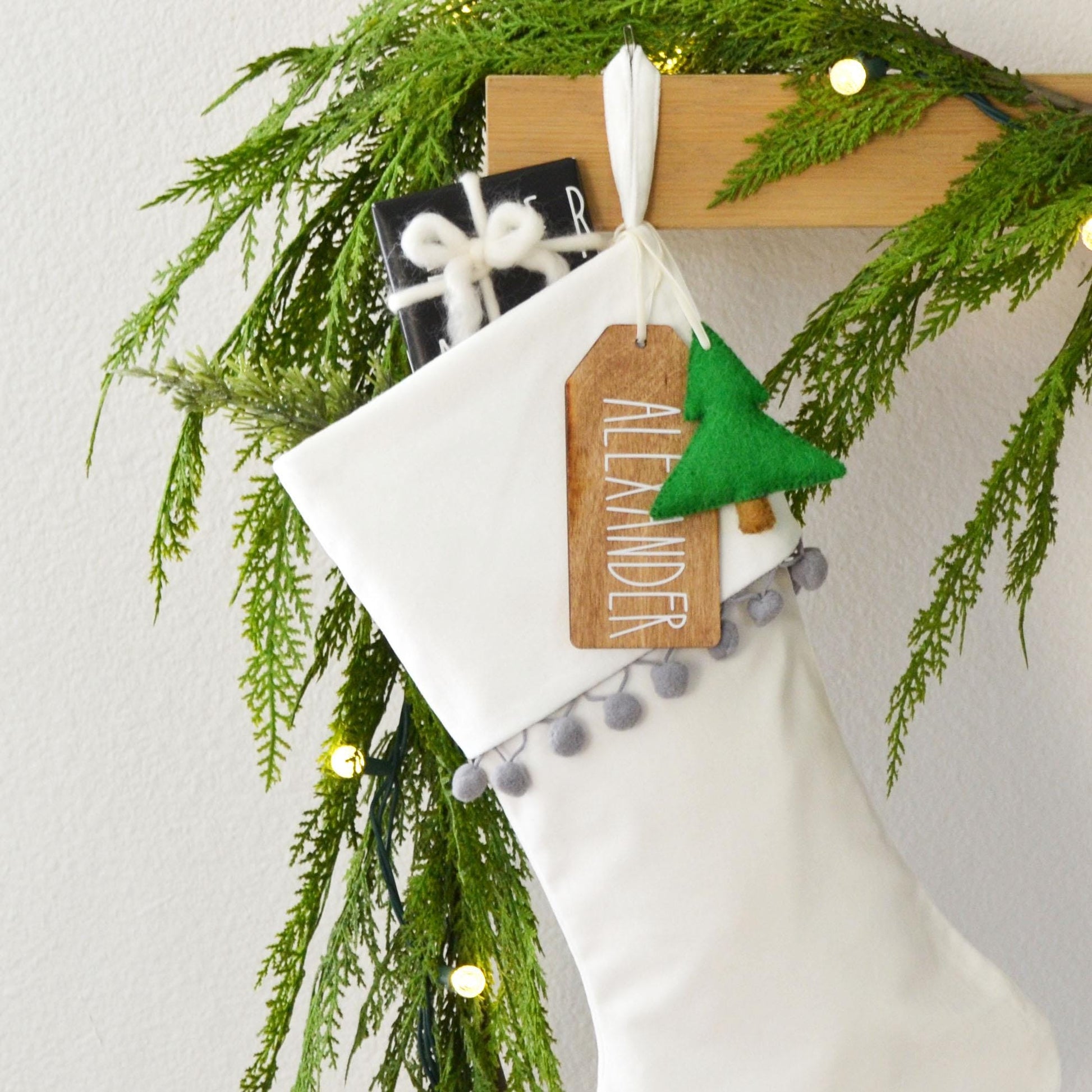 A white Christmas stocking decorated with greenery, a wooden tag, and a small green ornament, hanging on a wooden hanger.
